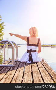 Young girl with white dress is sitting on a footbridge and enjoys the view over the lake.