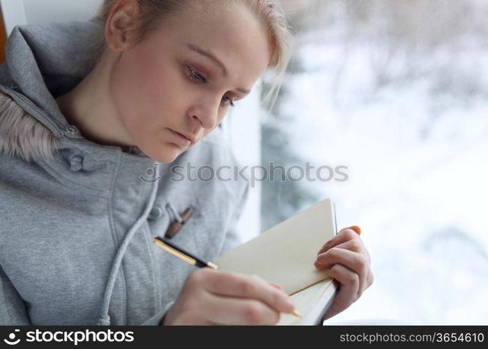 Young girl writing in her journal while sitting at a large window.