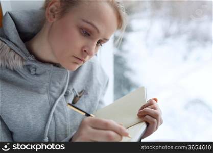 Young girl writing in her journal while sitting at a large window.