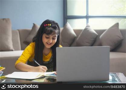 Young girl writing notes in her notebook while sitting in front of her laptop at home during online class