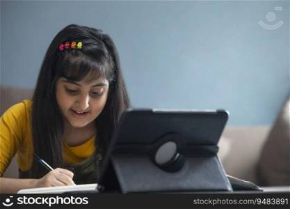 Young girl writing notes in her notebook while sitting in front of her Tablet at home during online class