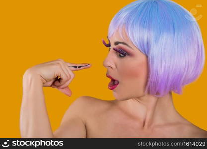 Young glam model in wig looking at mustache painted on finger looking excited on orange. . Playful model with mustache painted on finger