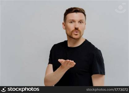 Young good looking unshaven man in basic black t shirt blowing sending air kiss to his girlfriend, trying to express love or flirt while standing isolated over grey studio background. Handsome guy sending air kiss and looking at camera