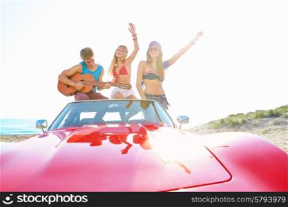 young group having fun on the beach playing guitar and dancing in a convertible car