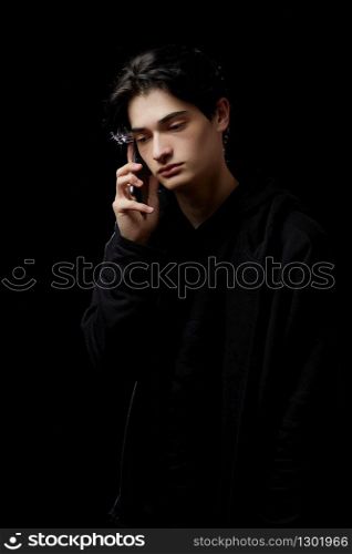 young guy in a black hoodie on a black background with a mobile phone in his hands