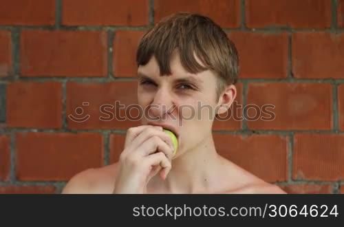 young guy sits in front of brick wall and appetite eats apple, closeup