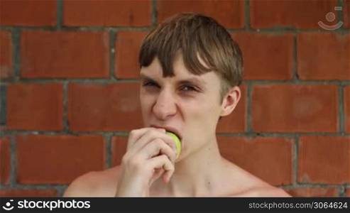 young guy sits in front of brick wall and appetite eats apple, closeup
