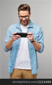 Young handcome man playing with portable video game standing over grey background.. Young handcome man playing with portable video game standing over grey background