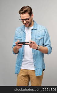 Young handcome man playing with portable video game standing over grey background.. Young handcome man playing with portable video game standing over grey background