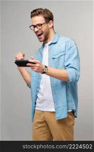 Young handcome man playing with portable video game standing over grey background.. Young handcome man playing with portable video game standing over grey background