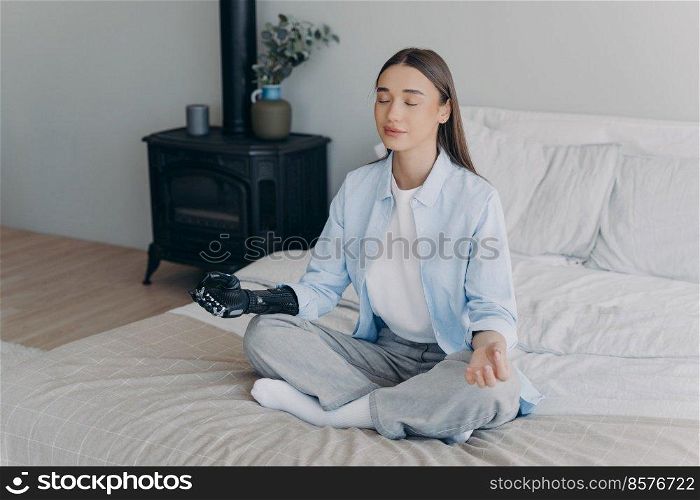 Young handicapped woman relaxing in cosy room. Girl practicing yoga in lotus pose on bed in her bedroom. Stress relief at home. Attractive european girl has cyber prosthesis. Modern artificial limb.. Young handicapped woman relaxing in cosy room. Girl practicing yoga in lotus pose on bed.