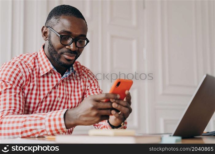 Young handsome african male executive in glasses sitting at office table and using mobile phone, american businessman wearing reading online news on smartphone while working on laptop computer. Young handsome african male executive in glasses sitting at office table and using mobile phone