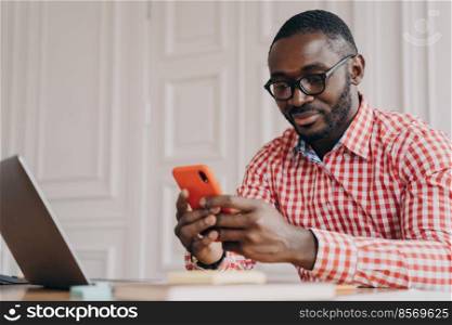 Young handsome african male executive in glasses sitting at office table and using mobile phone, american businessman wearing reading online news on smartphone while working on laptop computer. Young handsome african male executive in glasses sitting at office table and using mobile phone