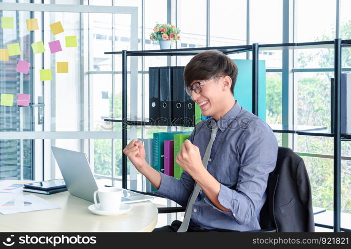 Young handsome asian businessman sitting at desk looking at labtop and raises hands with celebration impressive fiscal results or job success in meeting room. Business and successful concept