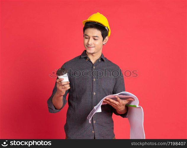 Young handsome asian man in casual dress with yellow hardhat hand holding paperwork blueprint and paper coffee cup in architect and engineer worker isolated in red background in studio.