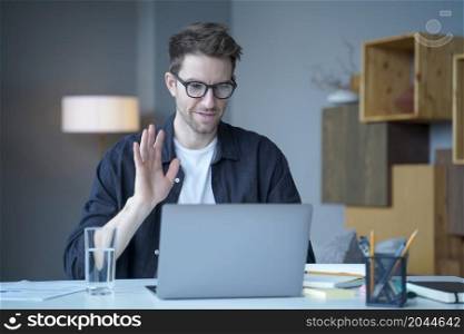 Young handsome austrian man freelancer in glasses waving hand in hello gesture during video call on laptop computer, male employee greeting colleagues at online meeting while working remotely at home. Young handsome austrian man freelancer waving hand in hello gesture during video call on laptop