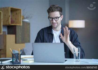 Young handsome austrian man freelancer in glasses waving hand in hello gesture during video call on laptop computer, male employee greeting colleagues at online meeting while working remotely at home. Young handsome austrian man freelancer waving hand in hello gesture during video call on laptop