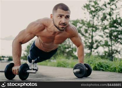 Young handsome bearded sportsman works out outdoor poses in plank with barbells, has strong body, muscular arms. Bodybuilder does exercises in open air, performs weight lifting, push up exercises