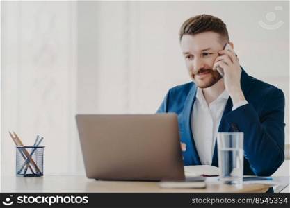 Young handsome business professional in suit talking on phone in front of laptop while listening attentively and checking information on laptop, sitting in stylish office with minimalistic design. Young businessman talking on phone in front of laptop behind office desk