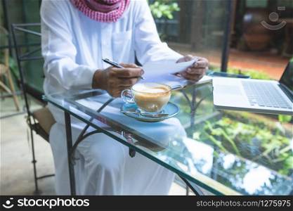 Young handsome businessman arab muslims in coffee shop wearing hijab dressed write on book file with laptop on table.focus on glass of hot coffee.