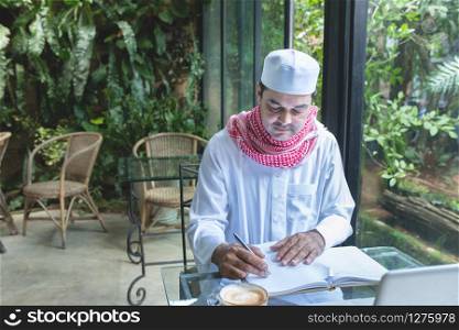 Young handsome businessman arab muslims in coffee shop wearing hijab dressed write on book file with laptop on table.