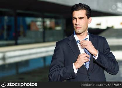 Young handsome businessman, dressed in blue suit, adjusting a tie in urban background with modern office buildings. Caucasian man with blue eyes.