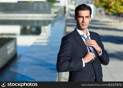 Young handsome businessman, dressed in blue suit, adjusting a tie in urban background with modern office buildings. Caucasian man with blue eyes.