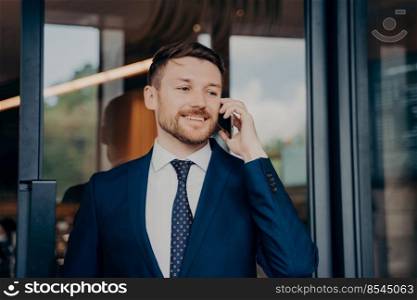 Young handsome businessman in suit talking on phone about important business, being satisfied by hearing good news about working process while standing alone in front of building entrance. Young businessman in blue suit talking on phone and smiling