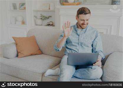 Young handsome casually dressed man waving his hand while speaking with family online using notebook, happy to have conversation with relatives, sitting on couch with leg crossed. Young man speaking with family using notebook while sitting on couch