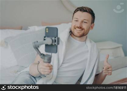Young handsome caucasian bearded man doing online call with webcam using smartphone while lying on bed at home, happy with big smile doing super cool sign thumb up with fingers. Young positive bearded man showing thumb up while doing online call on smartphone at home