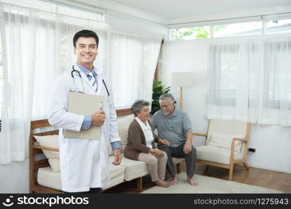 Young handsome caucasian male doctor standing hand holding document file look at camera with smile and two elderly old senior asian couple sit on couch in background,healthcare and medical concept.