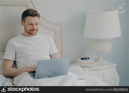 Young handsome cheerful smiling man freelancer in casual pajama outfit sitting in bed in morning and working remotely on laptop at home while resting in cozy bed. Freelance concept. Young handsome cheerful smiling man in casual pajama outfit sitting in bed and working on laptop