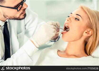Young handsome dentist examining teeth of happy woman patient sitting on dentist chair in dental clinic. Dentistry care concept.. Young dentist examining patient in dental clinic.