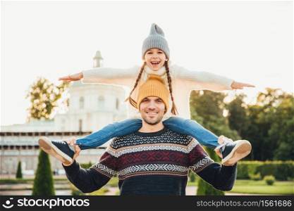 Young handsome father gives piggyback to his little smiling daughter, have fun together when have excursion. Small child looks at horizont while being at father`s back. Man gives girl ride on back