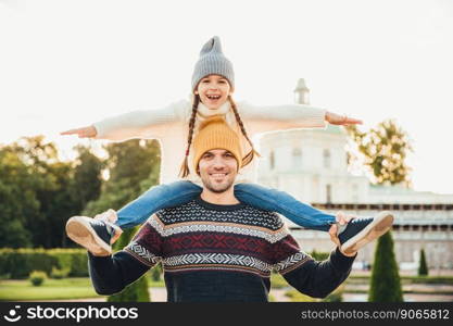Young handsome father gives piggyback to his little smiling daughter, have fun together when have excursion. Small child looks at horizont while being at father s back. Man gives girl ride on back