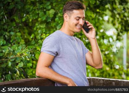 Young handsome looking guy talking with someone on phone. Man using his smartphone in park outdoor calling to somebody.. Young man talking on smartphone outdoor