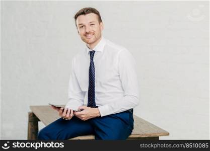 Young handsome male employee dressed elegantly, holds modern touch pad, thinks about financial report and meeting with business partners, sits at table, poses indoor. People and occupation concept