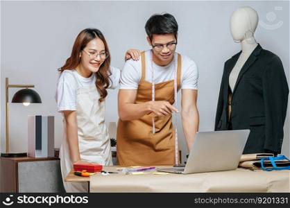 Young handsome man and pretty woman, Couple designer use laptop computer for searching good idea to design fashion clothing and chatting to customer online order, mannequin and tailoring tools on desk