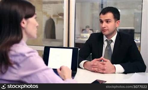 Young handsome man in corporate clothing sitting at the desk and taking about his successful work experience during job interview at the office. Young brunette female headhunter interviewing candidate to fill business position.