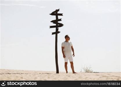 Young handsome man in light clothing and sunglasses near in the desert. column with plates to indicate the direction. Concept of freedom relaxation. Place for text or advertising.. Young handsome man in light clothing and sunglasses near in the desert. column with plates to indicate the direction. Concept of freedom relaxation. Place for text or advertising