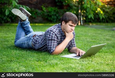 Young handsome man lying on grass at park and using laptop