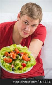 Young handsome man preparing to eat fresh healthy salad sitting on sofa at home and welcoming you to try too