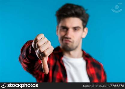 Young handsome man standing on blue studio background expressing discontent and showing thumb down gesture at camera. Portrait of guy with sign of dislike.. Young handsome man standing on blue studio background expressing discontent 
