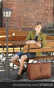 Young handsome man traveler casual style sitting on bench with suitcase waits, old town Gdansk Poland Europe