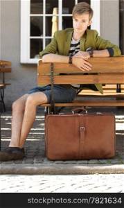 Young handsome man traveler casual style sitting on bench with suitcase waits, old town Gdansk Poland Europe