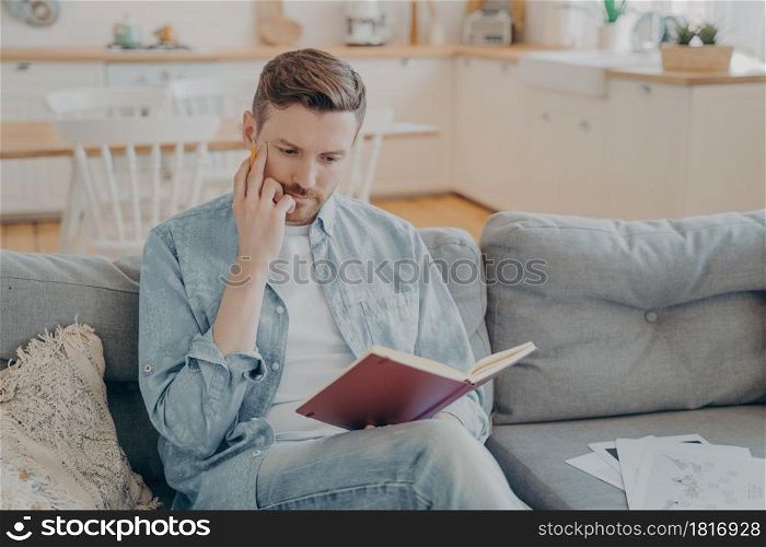 Young handsome man with hand on his face holding pencil, red note book on knee, making focused and serious gesture, tasked with finding new ideas, thinking what to write down. Young pensive handsome man finding new ideas and writing them down