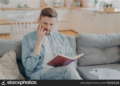 Young handsome man with hand on his face holding pencil, red note book on knee, making focused and serious gesture, tasked with finding new ideas, thinking what to write down. Young pensive handsome man finding new ideas and writing them down
