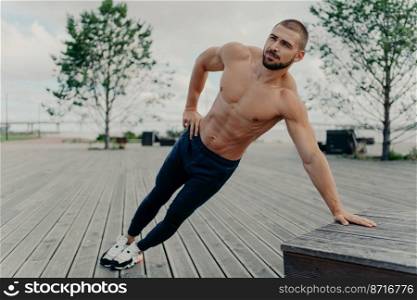 Young handsome shirtless ordinary bearded man stands in diagonal body position, makes side plank, practices endurance and stays in good physical shape. Sportsman in active wear trains outdoor