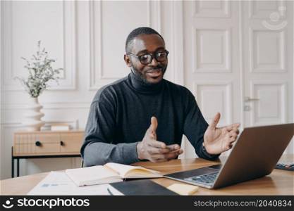 Young handsome smiling african american businessman in glasses enjoying video call on laptop computer, happy dark skinned male employee talking with colleague during online conference. Young smiling african american businessman in glasses enjoying video call on laptop computer
