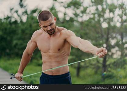Young handsome strong European man poses at park stadium, stretches skipping rope, has naked torso and muscular arms, going to have physical exercises, poses in open air. Street workout concept